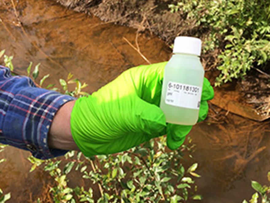 A gloved hand holding a sample tube above a creek. Photo.