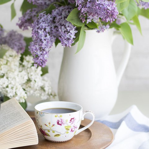 Lila syrener i en vit vas och en blommig kaffekopp, foto.
