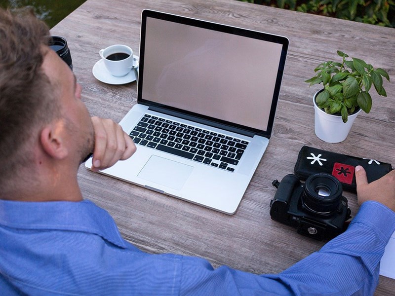 En man sitter vid ett bord utomhus med en kopp kaffe och en laptop