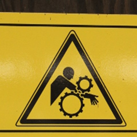 En person klämmer sin arm mellan två kugghjul, varningsskylt.