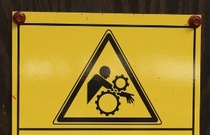 En person klämmer sin arm mellan två kugghjul, varningsskylt.
