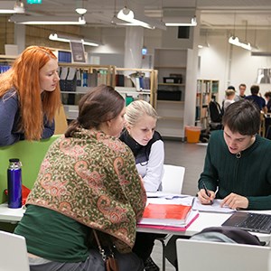 Four people are looking in a book togheter. Photo. SLU's library in Umeå. Photo.