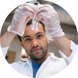 A man in a white lab coat holds up two test tubes. Photo.