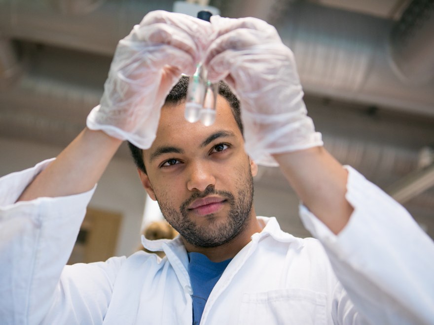 A man in a white lab coat holds up two test tubes. Photo.