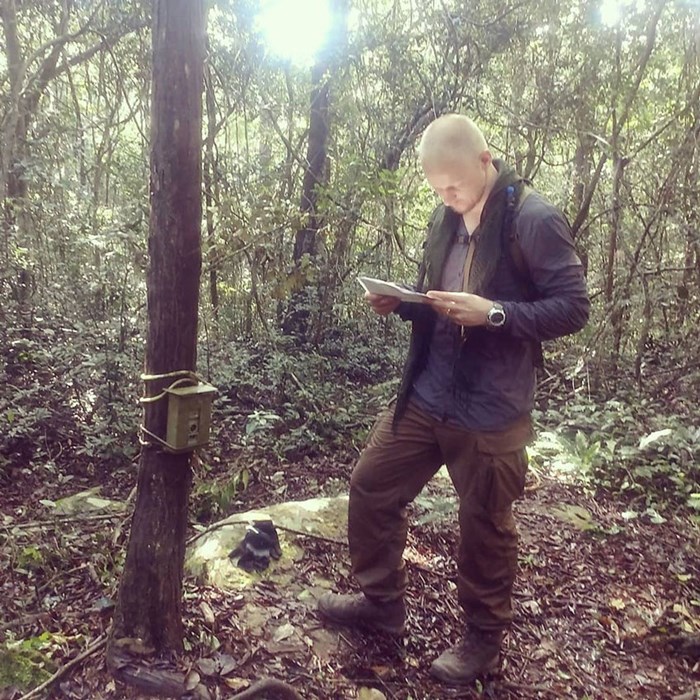 Adam Turner in a forest reading a paper. A camera trap is placed o one of the trees. Photo.