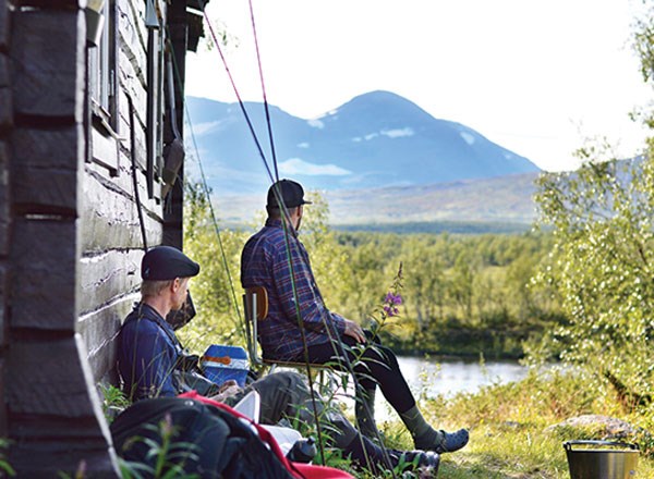 Två personer tar en paus vid en fjällstuga. Bredvid står fiskespön. Foto.
