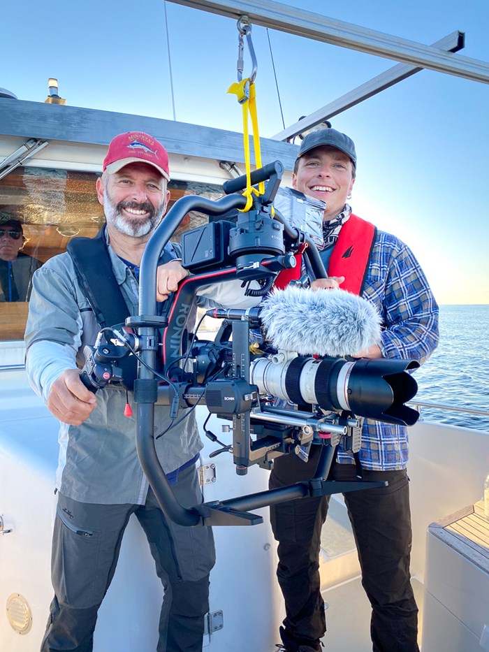 Två män bakom en stor filmkamera ombord på en båt. Foto.