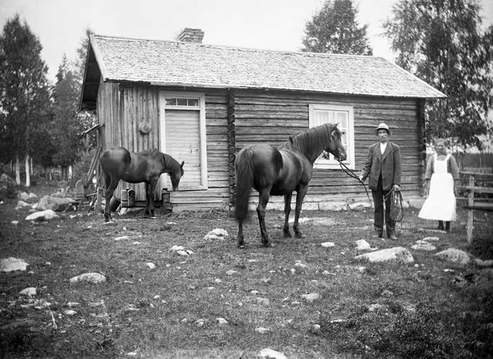 Äldre svartvitt fotografi av en man, kvinna och två hästar framför en byggnad.