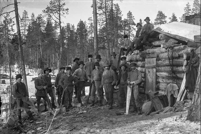 Äldre svartvitt fotografi av ett flertal män i skogen framför en timrad byggnad.
