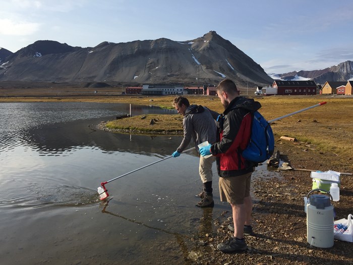 Two persons taking water samples from the shoreline. Mountains in the background. Photo.