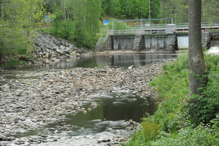 Dam gates, three closed and one open, with a stony river bottom in the front. Photo.