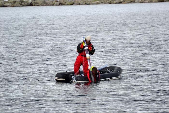 Two persons in a rubber boat. One of them is standing and is holding a long sediment tube. Photo.