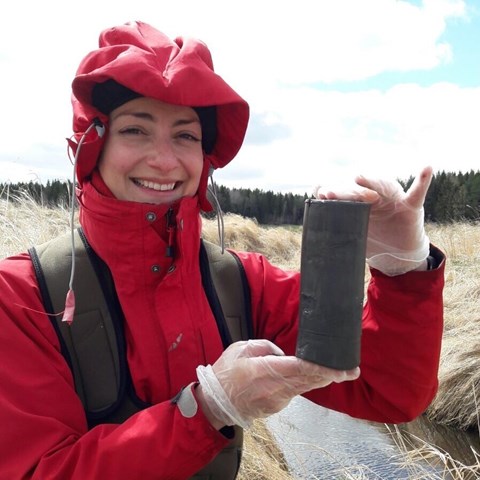 Person holding a compact sediment sample in front of a stream. Photo.
