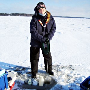Woman standing on an ice covered lake taking samples from a drilled hole. Photo.