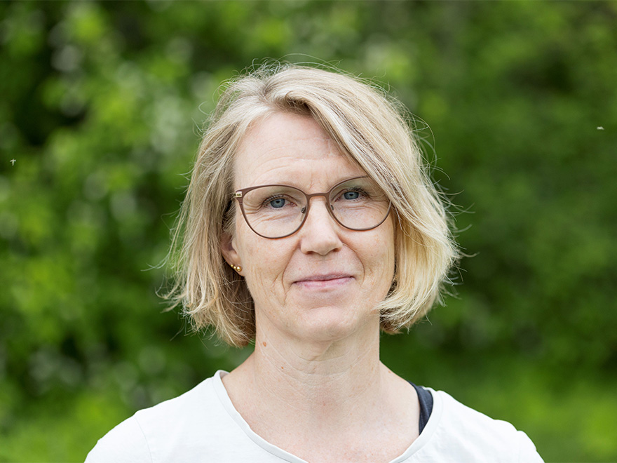 Kvinna med mellanblon page och glasögon tittar in i kameran. Foto