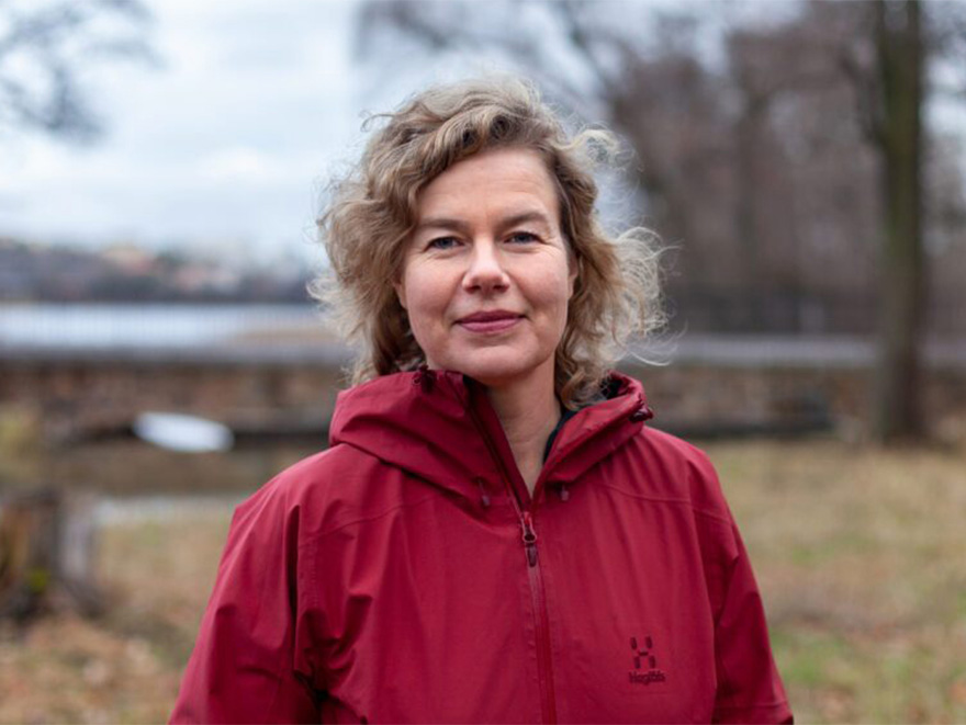 Kvinna med blond hår och röd jacka står i naturen och tittar mot kameran. Foto