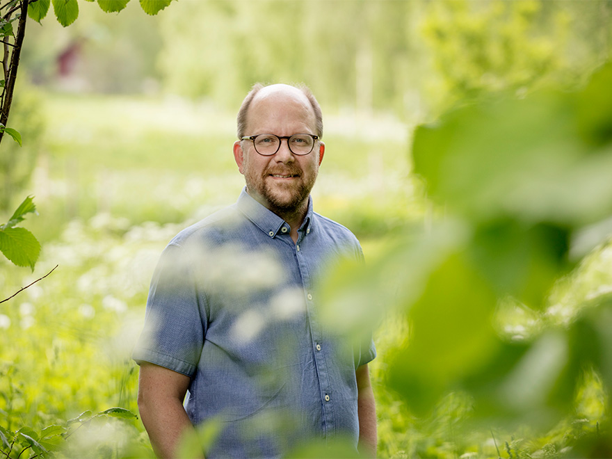 Man med blå skjorta står utomhus bland träd som grönskar. Foto