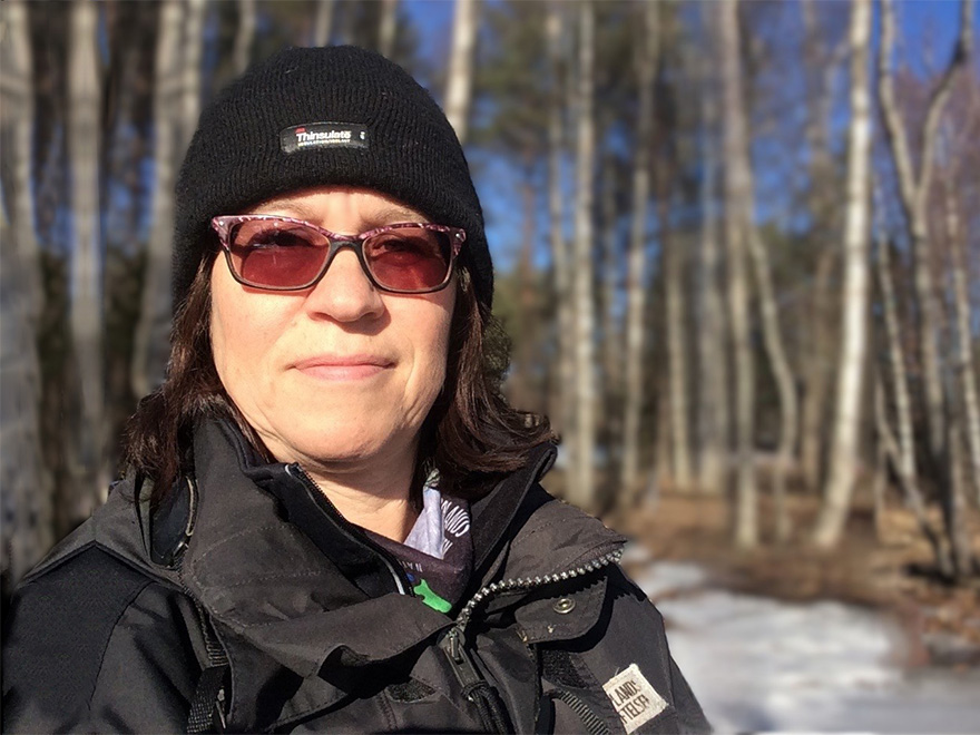 Kvinna med mörkt hår och tonade glasögon står i vintrig natur med solljus i ansiktet. Foto