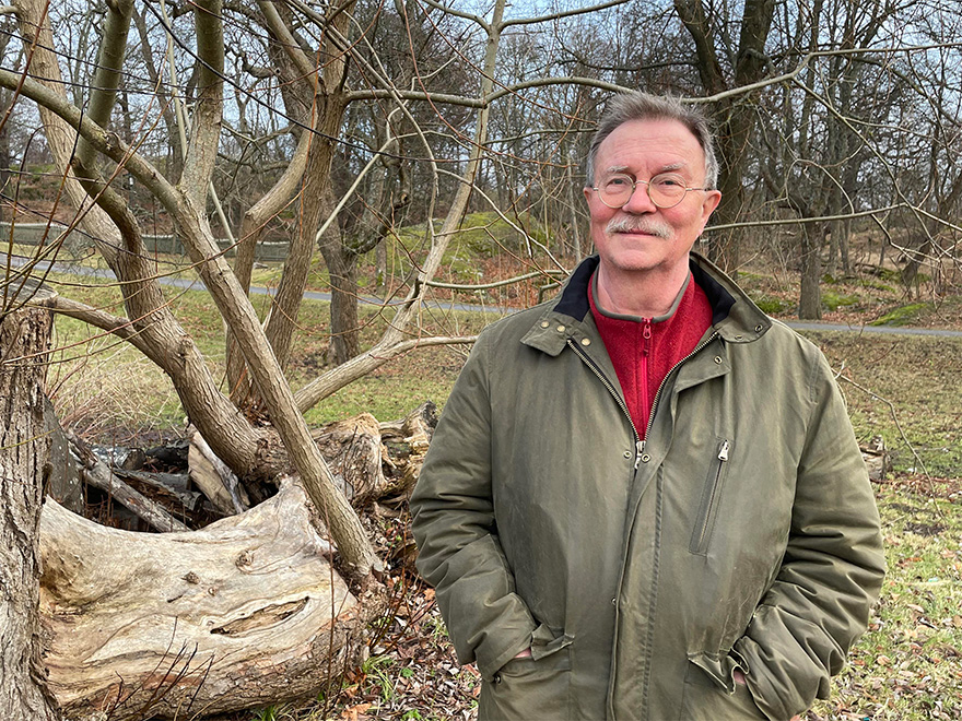 Man med grå mustasch och glasögon står med händerna i fickorna framför kala träd. Foto