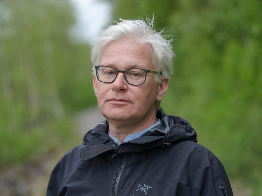 Man med vitt hår och mörk jacka står ute i naturen. Foto