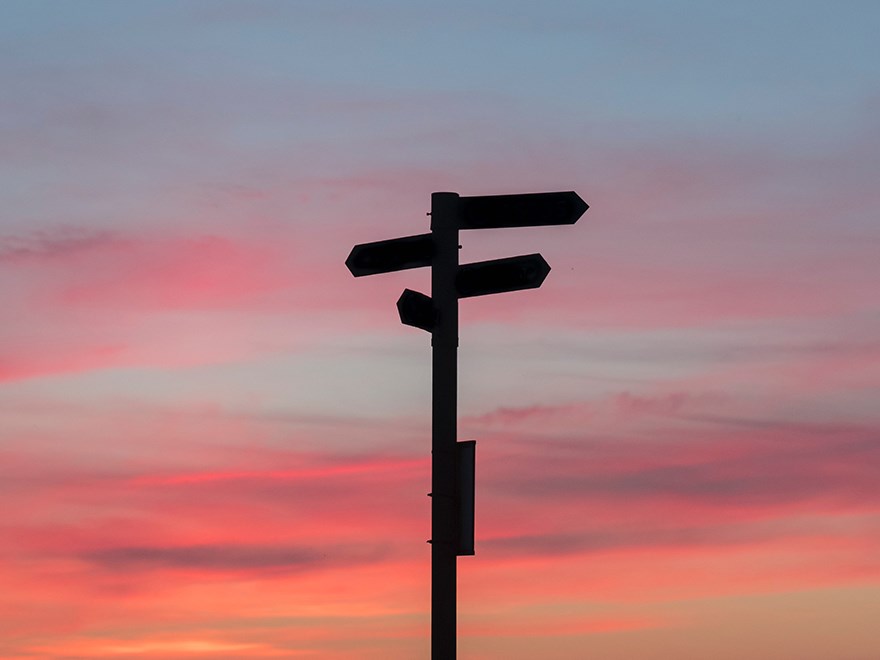 Med en rosa himmel i bakgrund visar fotot en stolpe pilar åt olika håll på. Foto. 