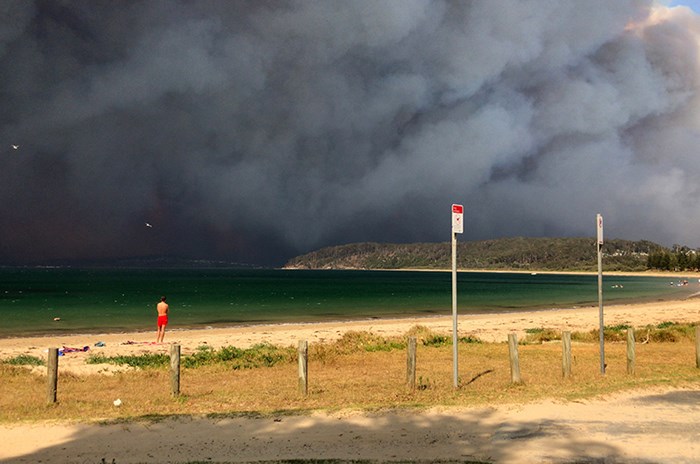 A beach by a sea, there is dark smoke all over the sky, photo.