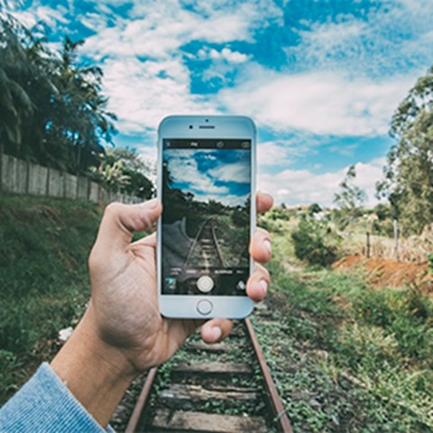 En hand håller i en mobiltelefon och fotar ett järnvägsspår, foto.