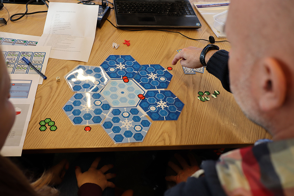 Two people are sitting at a table with a game board and paper in front of them. Photo.