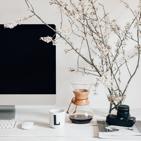 Ett skrivbord med en dator, tangentbord, mus, en kaffekanna och kaffekopp. På bordet finns även en bok, en kamera och en vas med kvistar med vita blommor. 