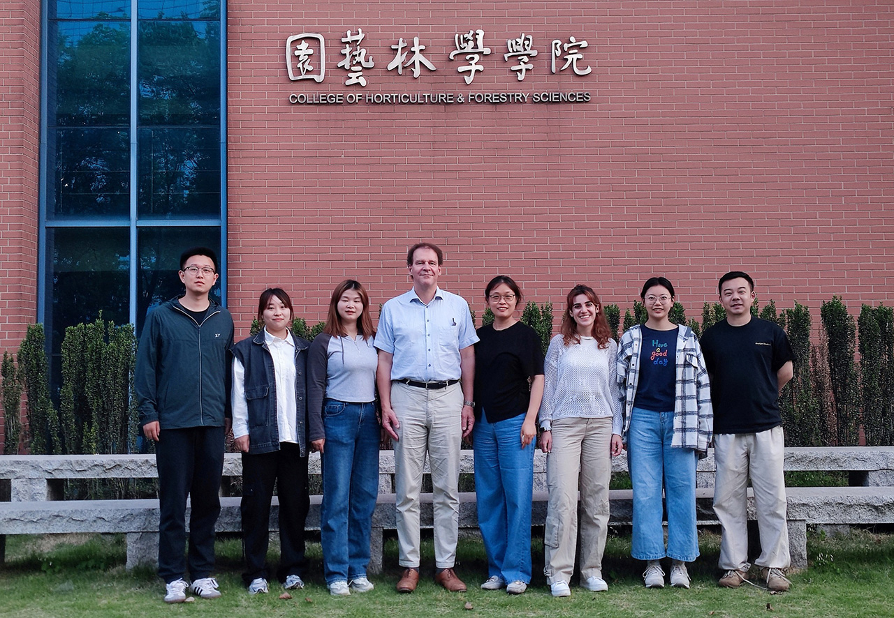 Åtta personer står uppradade bredvid varandra framför en röd tegelbyggnad.