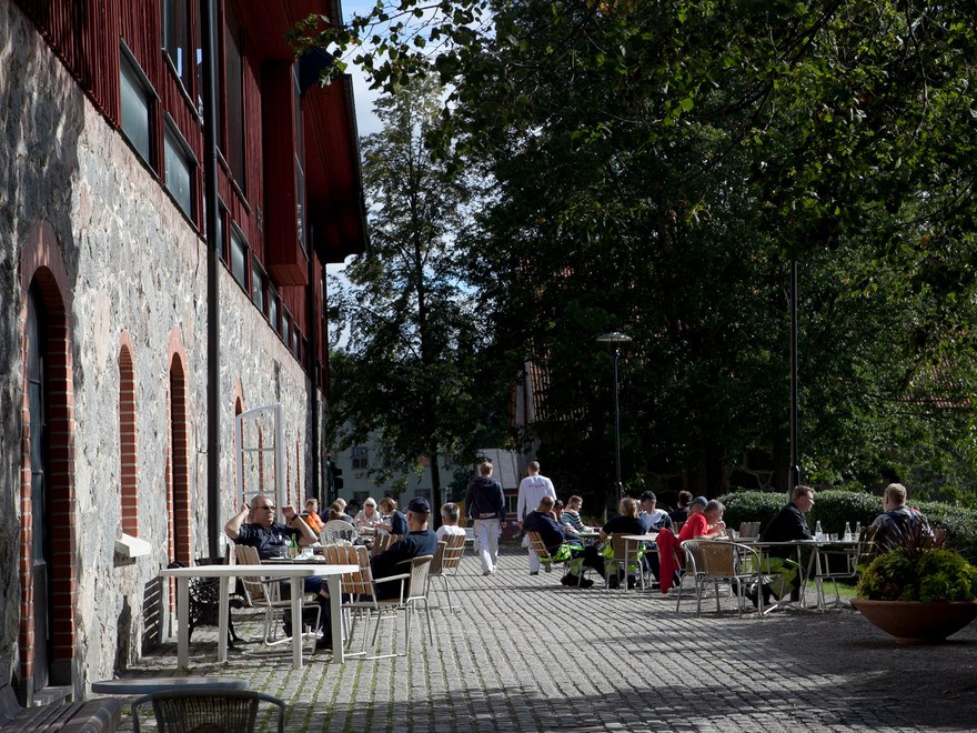 An image of people sitting outside. Summer, green trees on the right. A house made of stone and wood to the left.