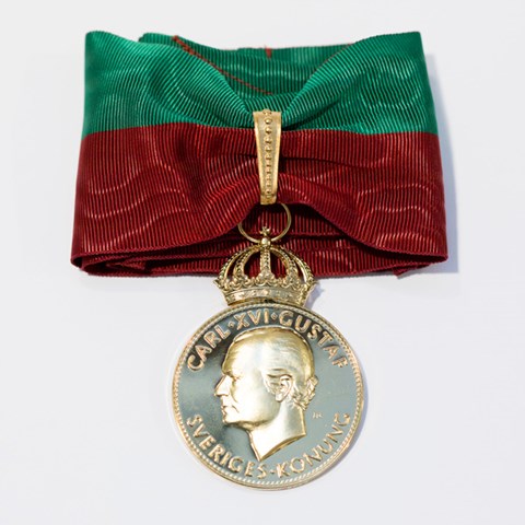 Bild på SLU:s stora förtjänstmedalj i guld. Medaljen är fäst på ett textilt band i universitetets färger rött och grönt. 