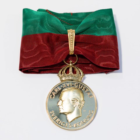 Bild på SLU:s stora förtjänstmedalj i guld. Medaljen är fäst på ett textilt band i universitetets färger rött och grönt.