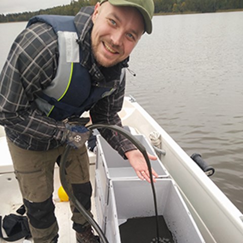 Oskar stands in a boat out on the lake and retrieves sediment samples through a hose. Photo.