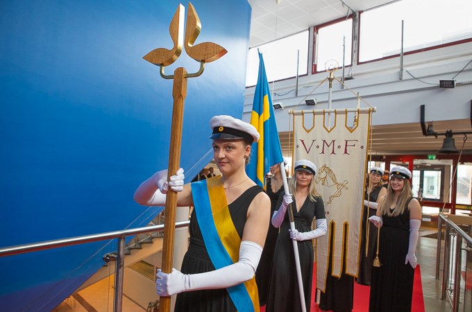 Bilden visar högtidsklädda studentmarskalkar med fanor. Kvinnan längst fram håller i SLU-staven. 