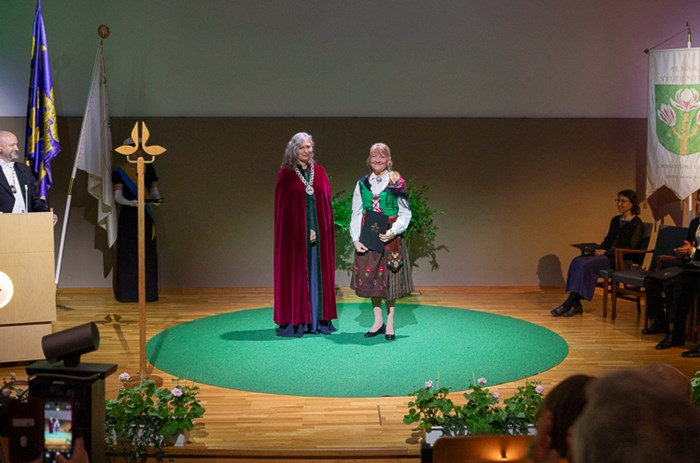 Två personer står på en scen på en grön matta, en med halskedja och röd sammetskåpa, den andra i färgglad folkdräkt.