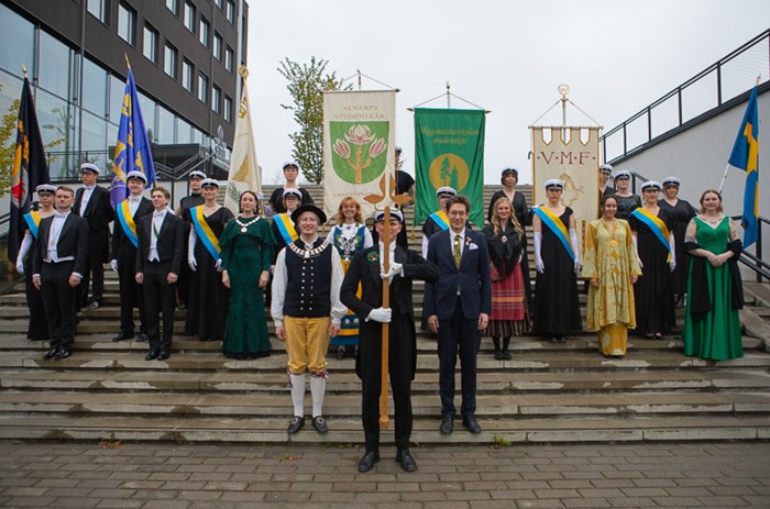 En grupp studenter i högtidskläder och studentmössor. En del av dem håller i fanor. 