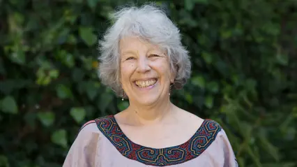 An older woman smiling in front of a bush.