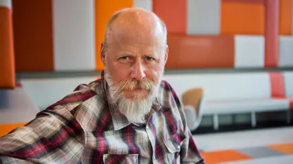 An elderly man with a white beard sitting in a modern indoor environment with orange coloured accents, looking firmly into the camera.