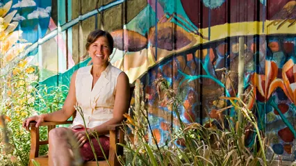A woman sitting outdoors in a wooden chair in front of a wall with graffiti.