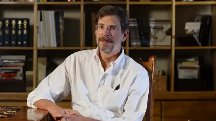 A man with dark hair wearing glasses and a white shirt sitting by a table in front of some bookshelves.