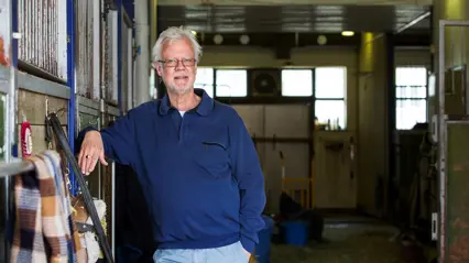 A man in a blue shirt leans himself against the wall in a stable.