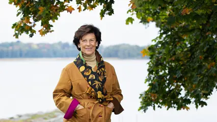 A woman in short brown hair and wearing a mustard yellow jacket is standing in an autumnal landscape, looking into the camera.