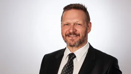 A smiling man with dark hair and beard, wearing a dark suit.