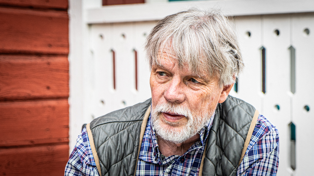 A man with grey hair and a grey beard looks out of the picture. In the background a red Swedish cottage.