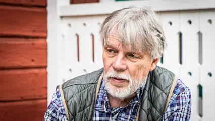 A man with grey hair and a grey beard looks out of the picture. In the background a red Swedish cottage.