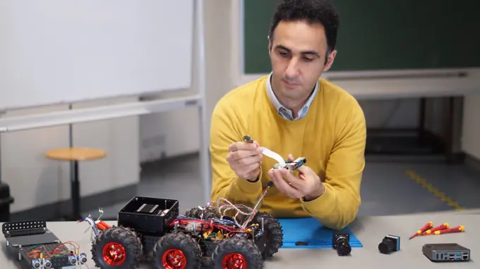 A man is sitting at a table assembling sensors and other electronic equipment onto a small wheeled robot.