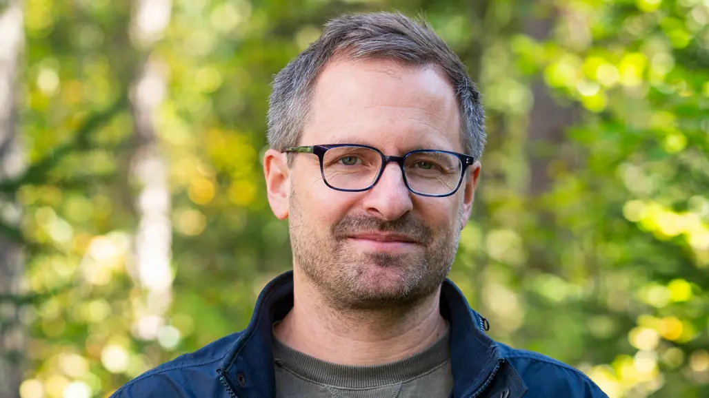 A man with short hair and glasses smiles into the camera, behind him blurry forest background.