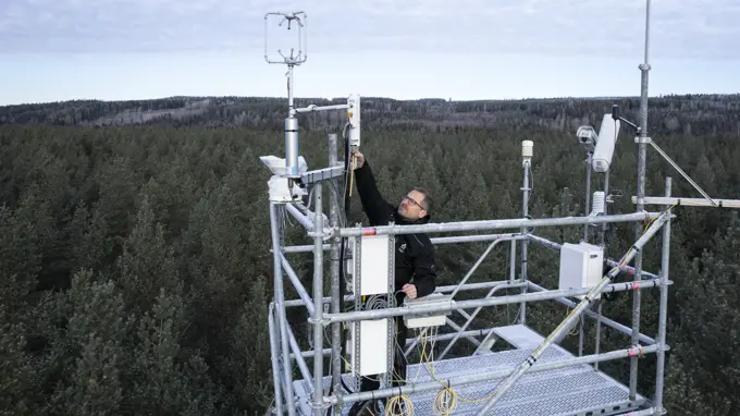 En forskare arbetar på en hög metallplattform mitt i en skog, omgiven av mätutrustning för att samla in miljödata.