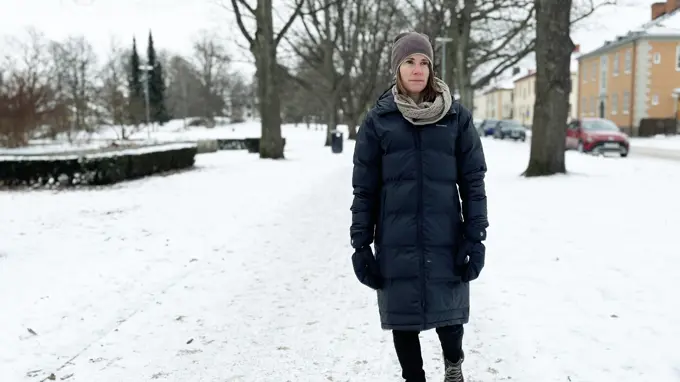 En kvinna går ensam genom en snötäckt park, klädd i vinterjacka, mössa och halsduk.